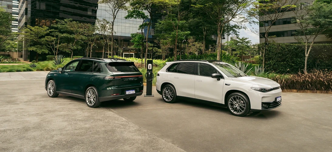 Dois SUVs Leapmotor, um verde escuro e outro branco, estacionados lado a lado em área urbana com prédios e árvores ao fundo.
