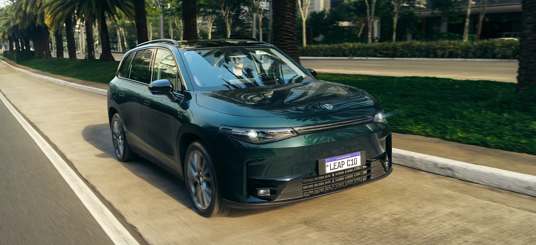 SUV Leapmotor verde escuro em movimento sobre via urbana, visto de frente, com faixa de rodagem, árvores e edifícios ao fundo.